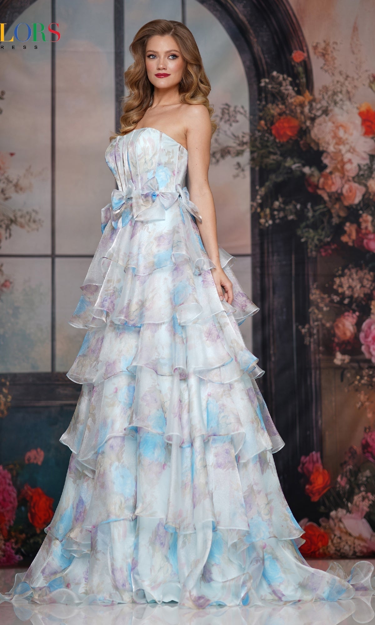 A woman wearing the Long Prom Dress: Colors Dress 3809, a strapless pastel tiered gown with a floral pattern and flowing chiffon A-line skirt, stands before large windows and colorful flowers.